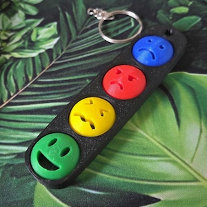 May include: A black keyring featuring four coloured smiley faces: green, yellow, red, and blue. The faces represent different emotions. The keyring has a silver ring for attaching keys.