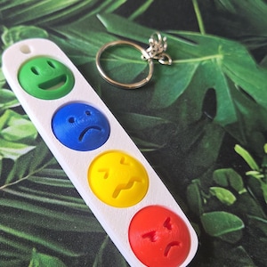 May include: A white rectangular keyring featuring four circular indentations, each containing a coloured emoji face: a green smiling face, a blue sad face, a yellow worried face, and a red angry face, accompanied by a silver metal split ring.