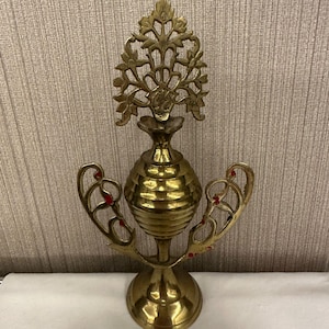 Puede incluir: Botella de perfume decorativa de latón con un diseño floral ornamental. La botella tiene un cuerpo bulboso y brazos curvos con detalles rojos. La base es redonda y el diseño general es intrincado. Altura aproximada 18 cm.