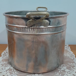 Puede incluir: Una olla de cobre grande de color plateado con dos asas de latón. La olla está sobre un encaje blanco.