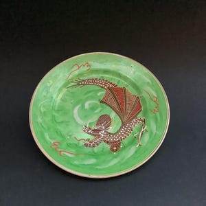 May include: A green ceramic plate featuring a red and brown dragon design. The dragon is shown in flight with its wings outstretched. The plate has a gold rim.