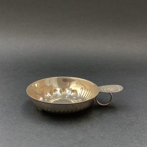 May include: A silver-toned metal tasting cup, known as a tastevin, featuring a shallow, circular bowl with a dimpled, reflective interior surface and a small, flat thumb rest handle with an engraved emblem and a circular finger ring attached.