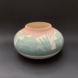 May include: A round, hand-painted ceramic pot with a pale pink rim and a turquoise body. The pot features white geometric designs and horizontal stripes. The interior is a light beige colour. The pot is set against a black background.