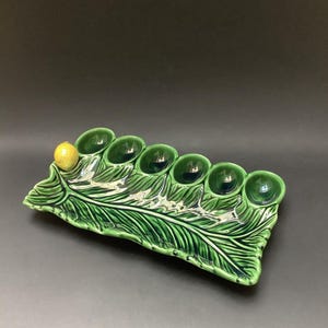 May include: A green ceramic egg holder shaped like a leaf, with six egg cups and a yellow lemon. The leaf design has detailed veining and a glossy finish. This decorative item is for the kitchen.