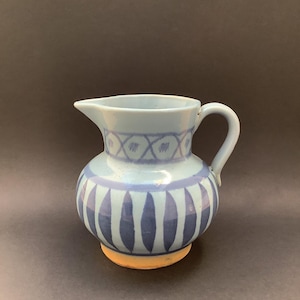 May include: A light blue ceramic jug featuring a dark blue painted pattern of vertical ovals around the base and a cross-hatch design around the neck. The jug has a curved handle, a pouring spout, and a visible unglazed orange-toned base.