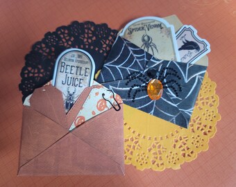May include: Halloween-themed craft featuring decorative envelopes, doilies, and labels. One envelope is brown, another is black with a spiderweb design and a jeweled spider. Labels read "Beetle Juice" and "Spider Venom".