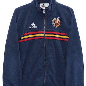 May include: Navy blue zip-up jacket with the Adidas logo and Spanish national team emblem. The jacket features red and yellow stripes across the chest. The jacket is hanging on a black hanger.