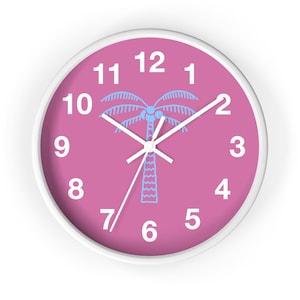 May include: Round wall clock with a white frame and a pink face. The clock face features white numerals, white hands, and a light blue palm tree graphic in the center. A decorative item for a home or office.
