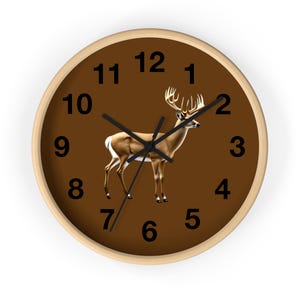 May include: A round wall clock with a brown face featuring a deer illustration. The clock has black numerals and hands, and a light wood frame. The deer is brown and white with large antlers.
