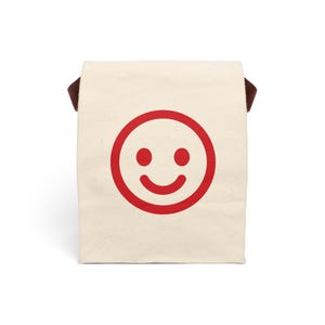 May include: A beige lunch bag with a large red smiley face graphic. The bag has a rectangular shape with a brown handle on each side. The smiley face features two red dots for eyes and a curved red line for a mouth.