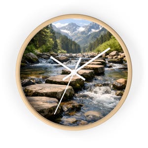 May include: Circular wall clock featuring a mountain stream scene. The clock face displays a photograph of a river with stepping stones, surrounded by trees and mountains. It has a light wood frame and white clock hands.