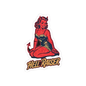 Op de afbeelding: Een sticker met een vintage-stijl illustratie van een roodhuidige vrouw met hoorns en een duivelsstaart, knielend en glimlachend. Ze draagt een donkerblauwe en gouden outfit. De tekst "Hell Raiser" staat eronder.