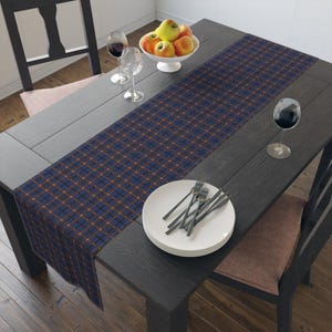 Puede incluir: Una mesa de comedor de madera oscura con un camino de mesa a cuadros azul marino y naranja. La mesa está puesta con platos blancos, cubiertos, copas de vino y un cuenco de manzanas. Una silla es visible a cada lado de la mesa.
