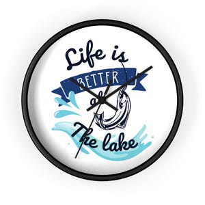 Puede incluir: Un reloj de pared redondo blanco con agujas negras y marco negro. La esfera del reloj presenta un pez azul saltando fuera del agua con el texto "Life is better at the lake".