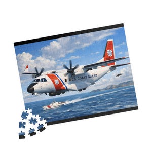 Puede incluir: Un rompecabezas que representa un avión de la Guardia Costera de EE. UU. y un guardacostas en el agua. El avión es blanco con detalles rojos y azules, y el texto "U.S. COAST GUARD" es visible. El rompecabezas está parcialmente montado.