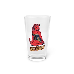 May include: A clear pint glass featuring a retro illustration of a red devil woman with horns and a tail. The text "Hell Raiser" is printed in yellow below the image. The glass is empty and sits on a white surface.