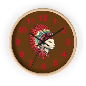 May include: A round wall clock with a brown face and a light wood frame. The clock features a white cat wearing a Native American headdress. Red numbers mark the hours.