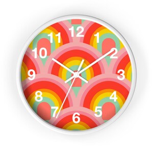 May include: A round wall clock with a white frame. The clock face features a repeating rainbow pattern in shades of red, orange, yellow, and green on a pink background. White hands and numerals complete the design.