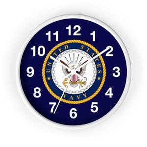 May include: A round wall clock with a white frame and a dark blue face featuring the United States Navy emblem in the center. The emblem displays a bald eagle, an anchor, and a chain border. White numbers from 1 to 12 mark the hours.