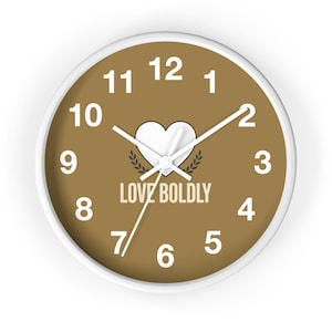 Op de afbeelding: Een ronde wandklok met een witte rand en een beige wijzerplaat. De klok heeft witte cijfers van 1 tot 12, witte uur-, minuut- en secondewijzers, een wit harticoon boven de tekst LOVE BOLDLY en kleine decoratieve takjes met blaadjes.