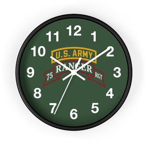Könnte beinhalten: Eine runde Wanduhr mit schwarzem Rahmen und dunkelgrünem Zifferblatt. Das Zifferblatt zeigt weiße Zahlen von 1 bis 12, weiße Zeiger und ein zentrales Motiv mit dem Text U.S. ARMY RANGER 75 RANGER RGT auf einem gelb-roten Bannerdesign.