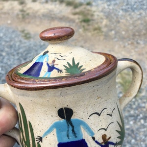 May include: Hand-painted ceramic teapot with a lid. The teapot has a beige speckled background with brown trim and a brown lid. The design includes figures in blue and brown, green cacti, and flying birds. The handle is curved.