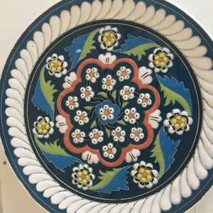 Puede incluir: Plato de cerámica decorativo con fondo verde azulado oscuro y diseño floral. El plato presenta un borde festoneado blanco, un patrón floral central con detalles en naranja, amarillo y azul, y hojas verdes.
