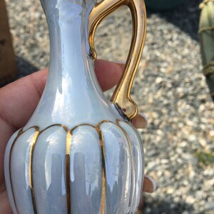 Puede incluir: Una pequeña jarra de cerámica azul claro con asa dorada y ribetes dorados. La jarra tiene un diseño acanalado y un borde ornamentado. Los detalles dorados contrastan con el color azul pálido.