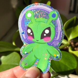 Puede incluir: Un extraterrestre verde con ojos grandes y una diadema morada con un acento azul. El extraterrestre lleva auriculares y tiene un fondo hologr&aacute;fico brillante.