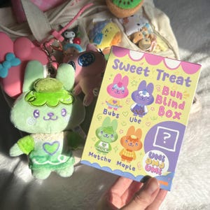 Op de afbeelding: Een lichtgroene pluche konijn sleutelhanger met een bijpassende outfit en een "Sweet Treat Bun Blind Box" met konijn karakters. De doos heeft de woorden "Bubs", "Ube", "Matcha" en "Maple" erop.