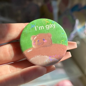 May include: A green button with a holographic finish features a cartoon brown bear with the text "I'm gay" in white.