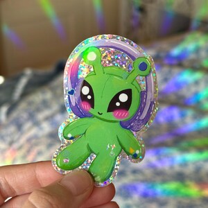 Puede incluir: Un extraterrestre verde con ojos grandes y un halo morado, que lleva una pegatina hologr&aacute;fica brillante. La pegatina tiene un acabado brillante y es perfecta para agregar un toque de fantas&iacute;a a cualquier superficie.