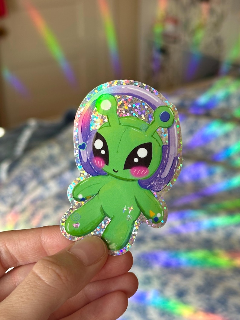 Puede incluir: Un extraterrestre verde con ojos grandes y un halo morado, que lleva una pegatina hologr&aacute;fica brillante. La pegatina tiene un acabado brillante y es perfecta para agregar un toque de fantas&iacute;a a cualquier superficie.