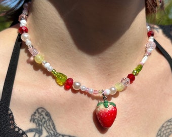 May include: A colorful beaded necklace with a strawberry charm. The necklace features white, pink, green, and red beads.