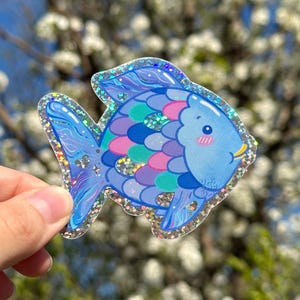 May include: A blue and purple glittery fish sticker with pink and green scales. The fish has a cute face with rosy cheeks and a small mouth.