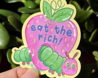 May include: A pink strawberry with green leaves and a caterpillar eating it. The strawberry has the text "eat the rich!" on it.