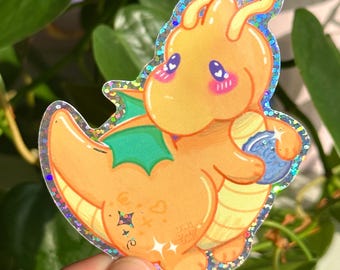 Puede incluir: Una pegatina holográfica con un dragón de dibujos animados en tonos naranja, amarillo y verde. El dragón tiene ojos grandes y sostiene un orbe azul. La pegatina tiene un borde brillante. Fondo de hojas verdes.