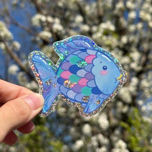 May include: A blue and purple glitter fish sticker with a pink and green scale pattern. The fish has a cute face with blush and a smile.