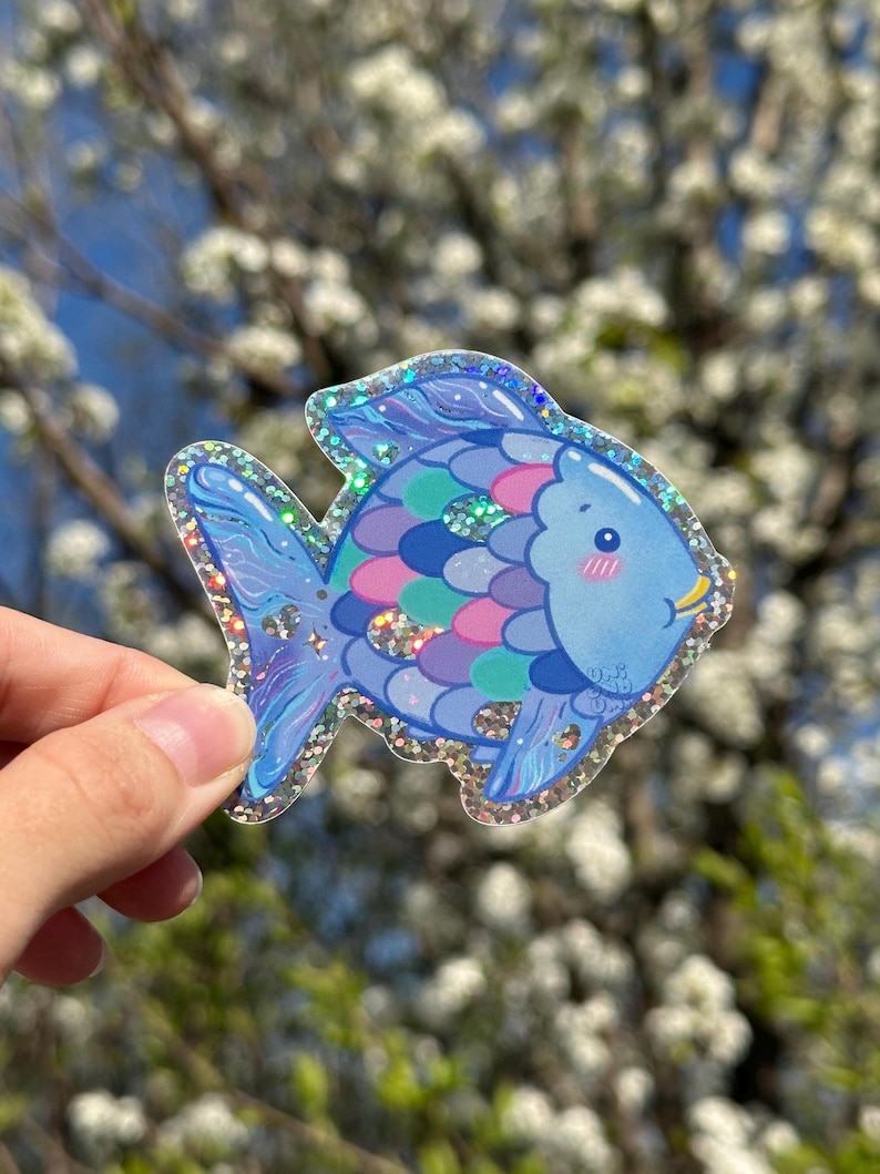 May include: A blue and purple glitter fish sticker with a pink and green scale pattern. The fish has a cute face with blush and a smile.