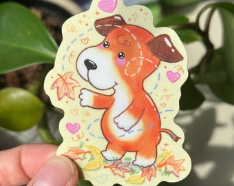Op de afbeelding: Een sticker met een cartoonhond met oranje vacht, bruine oren en een roze neus. De hond staat op een gele achtergrond met verspreide bruine bladeren en roze harten. De hond kijkt omhoog en heeft een vrolijke uitdrukking.