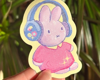 May include: A pink glitter bunny wearing blue headphones with colorful designs. The bunny is wearing a pink dress with glitter accents.