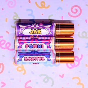 May include: Three perfume roller bottles with gold caps. Each bottle has a label with a circus tent design and a name: Jax, Pomni, and Ragatha. The background is a light purple with colourful confetti.