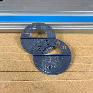 Puede incluir: Dos guías de medición de plástico gris con el texto "KERF Side" y "2 mm" impreso en ellas.