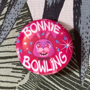 May include: A round, pink button with the words "BONNIE BOWLING" in white. The button features a cartoon image of a purple bunny with red eyes and a smiling mouth, surrounded by blue starbursts.