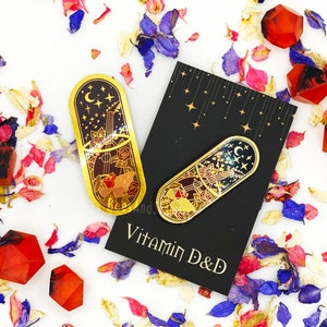 May include: Two gold-toned enamel pins with a black and red design. The pins feature a crescent moon, stars, and a guitar with roses. The pins are on a black background with gold stars. The text "Vitamin D&D" is on the background.