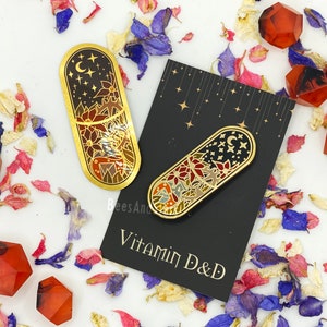 May include: Two gold enamel pins with celestial and floral designs, one larger and one smaller, displayed on a black card with the text "Vitamin D&D". The pins feature a night sky with stars and a crescent moon.
