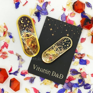 May include: Two gold-toned enamel pins shaped like capsules with a black background and a design of a deer with antlers, a crescent moon, and stars. The text "Vitamin D&D" is printed on a black card.