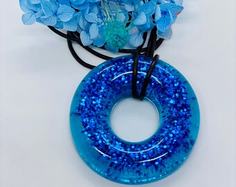 Puede incluir: Un colgante de donut de resina azul con purpurina y un cordón negro. El colgante está rodeado de flores azules y turquesas.
