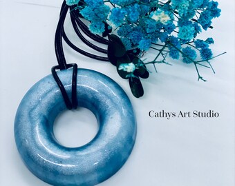 Puede incluir: Un colgante de dona de resina azul claro con un acabado de purpurina plateada. El colgante cuelga de un cordón de cuero negro. El colgante está rodeado de un ramo de aliento de bebé azul. El texto "Cathys Art Studio" es visible en la esquina inferior derecha de la imagen.