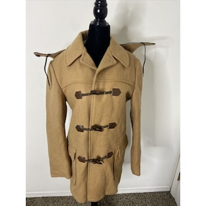 Könnte beinhalten: Ein beigefarbener Dufflecoat aus Wolle mit Kapuze und Knebelverschlüssen. Der Mantel hat ein klassisches Design mit zwei Vordertaschen und einem geknöpften Kragen. Die Knebelverschlüsse sind aus braunem Leder und Holz. Der Mantel wird auf einer schwarzen Schaufensterpuppe präsentiert.